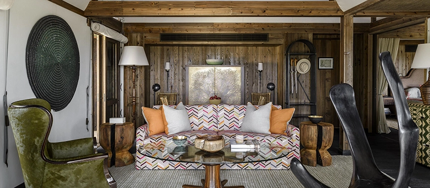 The living area of a Xigera suite, featuring timber-panelled walls, a chevron-patterned sofa with orange cushions, a large woven wall disc, and a sculptural dark wooden chair in the foreground.
