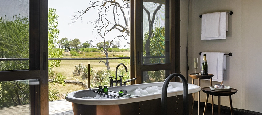 A copper freestanding bathtub positioned before floor-to-ceiling windows looking out over the green floodplains of the Okavango Delta, with a champagne bottle and glassware on a side table.