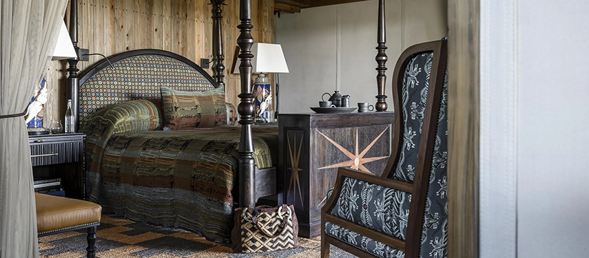 The bedroom of a Xigera suite, centred on a four-poster bed with carved dark wooden posts and richly layered patterned bedding, beside a high-backed chair upholstered in deep floral fabric.