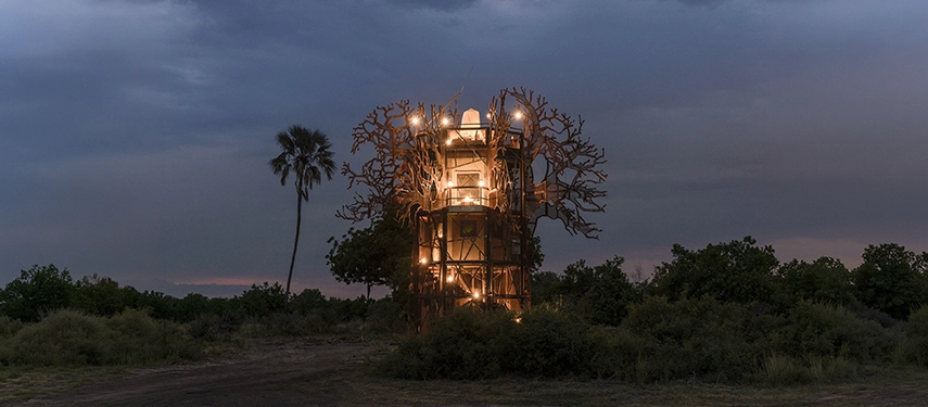The Xigera Baobab Suite illuminated at dusk, its sculptural steel framework evoking the spreading canopy of a baobab tree, glowing warmly against a darkening Delta sky.