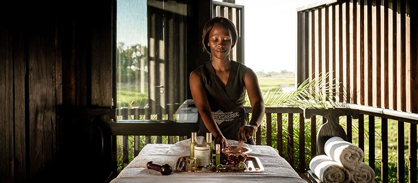 A Xigera spa therapist preparing a treatment table on an open-air deck, with the light-filled expanse of the Okavango Delta visible through slatted wooden screens behind her.