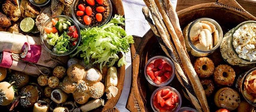 An overhead view of a bush picnic spread — bread rolls, salads in glass jars, cherry tomatoes, strawberries, breadsticks and petit fours arranged in wooden bowls on a white cloth.