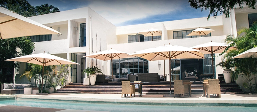 The pool and terrace facade of Latitude 15° in Lusaka, a crisp white modernist building with stepped outdoor seating, large parasols and a foreground of tropical garden planting.