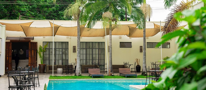 The pool area of The Other Side at Latitude 15° in Lusaka, with sun loungers, outdoor dining chairs and sail shades strung between palm trees against a backdrop of tropical greenery.