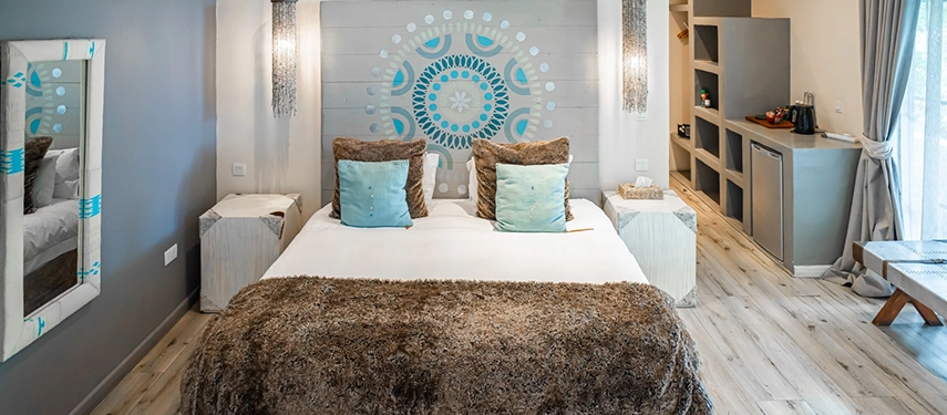 A guest room at Latitude 15° in Lusaka with light timber floors, a king bed with a fluffy brown throw and teal cushions, crystal wall sconces flanking a painted mandala headboard, and open shelving with a minibar.