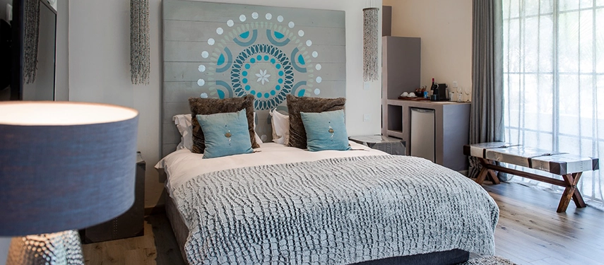 A spacious guest room at Latitude 15° in Lusaka with light timber floors, a king bed dressed in a textured grey bedspread and teal cushions, a painted mandala headboard, and large garden-facing windows flooding the room with natural light.