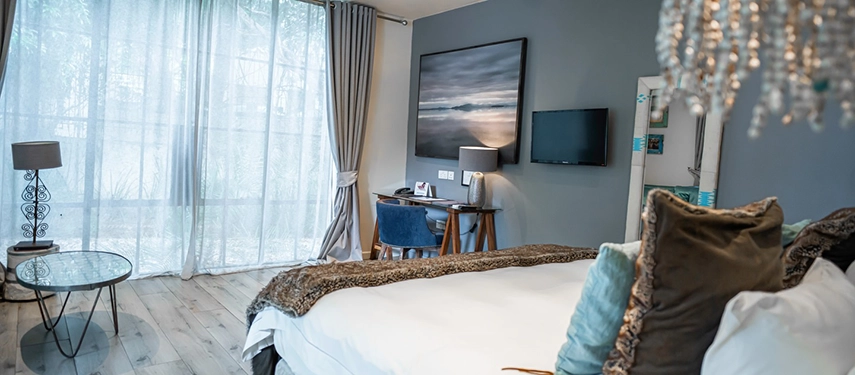 A guest room at Latitude 15° in Lusaka featuring a king bed with a rich brown throw, a crystal chandelier, floor-to-ceiling sheer curtains, a wall-mounted flat-screen television and a writing desk with blue upholstered chairs.