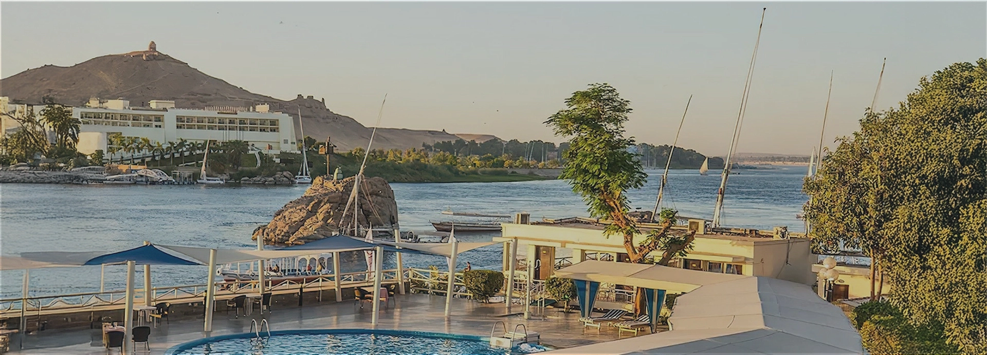 A sweeping view over the hotel pool and the Nile at sunset, with feluccas drifting past and desert hills beyond.