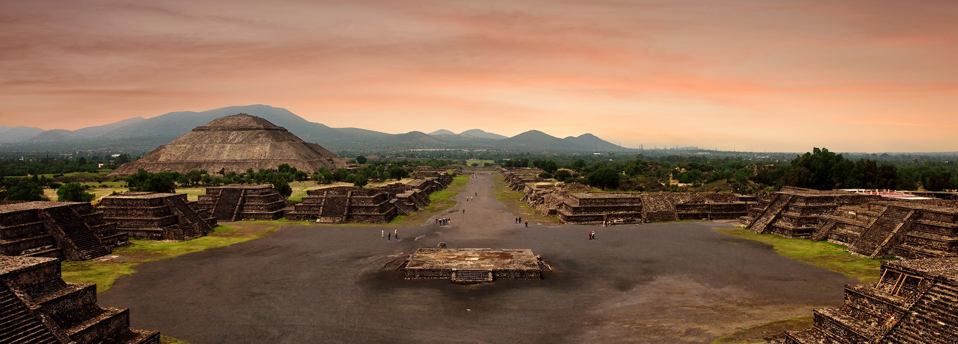 Looking along the Avenue of the Dead at Teotihuacán at dusk, the Pyramid of the Sun rising on the left and smaller temple platforms flanking the broad ceremonial boulevard beneath a warm pink and amber sky.
