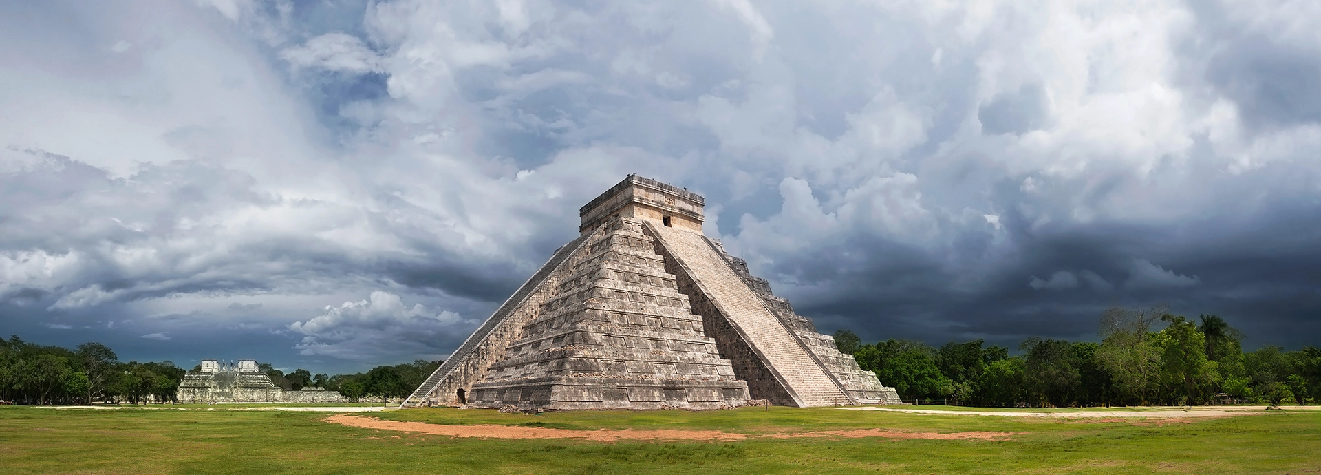 El Castillo pyramid at Chichén Itzá standing in dramatic relief against a brooding, cloud-filled sky, its stepped stone tiers and central staircase rising from an open grass plaza.