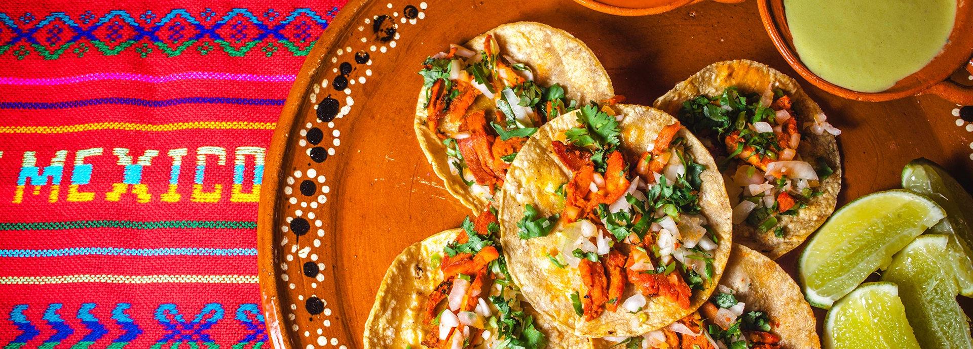 A hand-painted terracotta platter of tacos topped with spiced filling, diced onion and fresh coriander, served alongside lime wedges and green salsa on a colourful embroidered Mexican textile.
