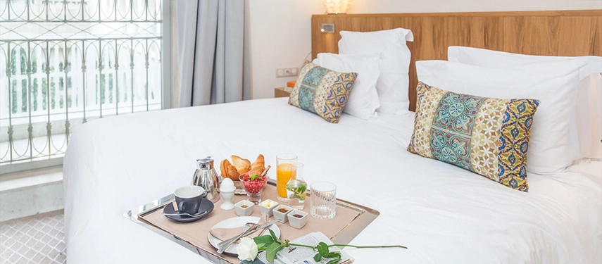 Inviting bedroom with breakfast tray on a crisp white bed, patterned cushions, and natural light from tall windows.