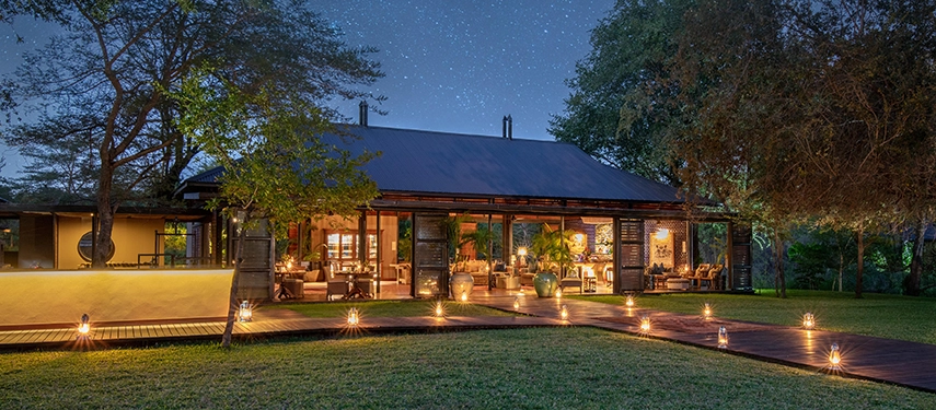 The open lounge and dining spaces at Mukwa River Lodge are styled with earthy tones, lantern light, and relaxed seating overlooking the gardens.