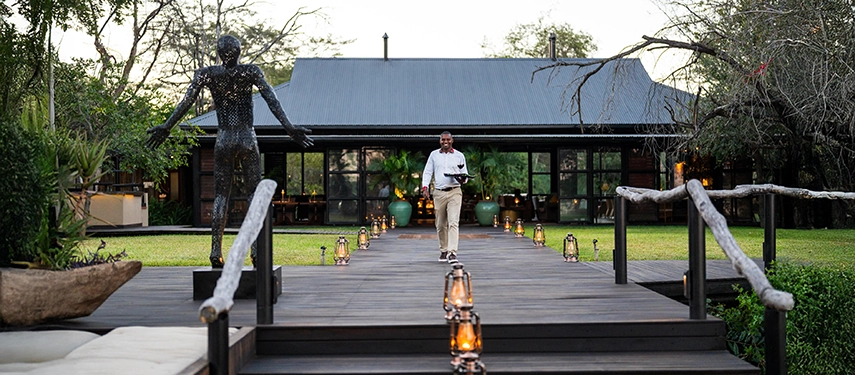 A lantern-lit walkway leads towards Mukwa River Lodge’s main building, framed by sculptures, soft lighting, and lush riverine gardens at dusk.