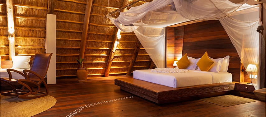 A serene bedroom scene with a handcrafted wooden bed, flowing mosquito netting and warm ambient lighting beneath a thatched roof.