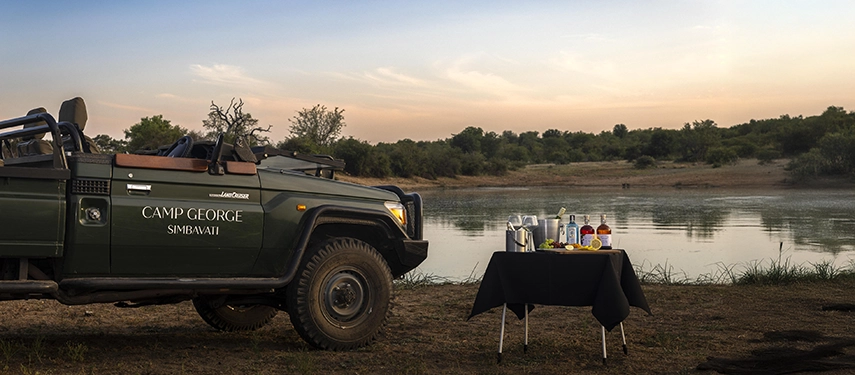 A classic safari vehicle is parked beside a river for sunset drinks at Simbavati Camp George, set against calm water and open skies.