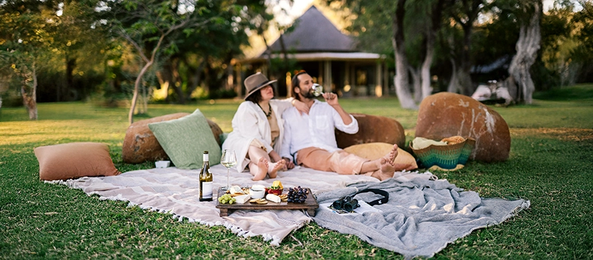 Guests relax on a picnic blanket at Simbavati Camp George, enjoying wine and local flavours in a peaceful garden setting.