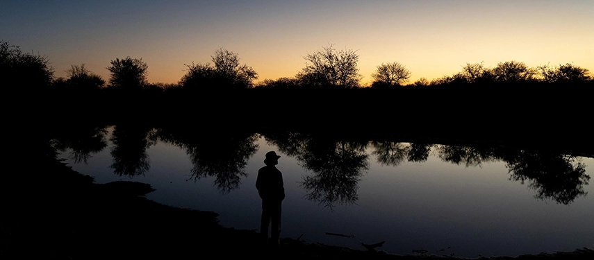 A silhouetted figure stands beside a still waterhole at sunset near Simbavati Camp George, capturing the quiet atmosphere of the Timbavati wilderness.