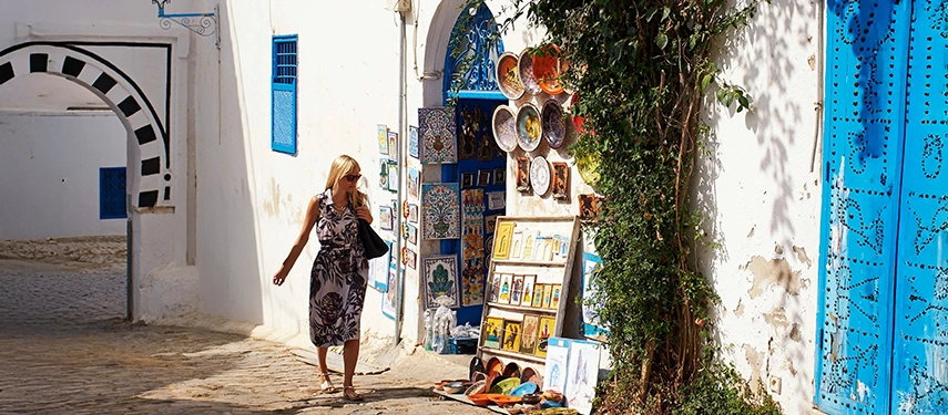 Woman walking past a vibrant artisan stall selling ceramics and paintings in the whitewashed streets of Sidi Bou Said, Tunisia.