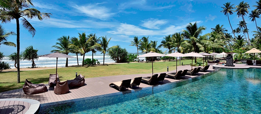 A relaxing poolside scene at Weligama Bay Marriott Resort & Spa with cushioned loungers, palm trees, and waves rolling onto the nearby shore.