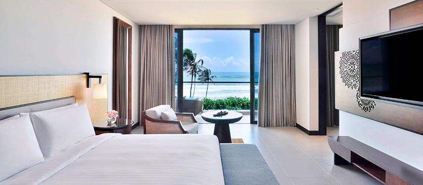 A bright, modern suite at Weligama Bay Marriott Resort & Spa with floor-to-ceiling windows framing palm trees and waves rolling onto the shore.
