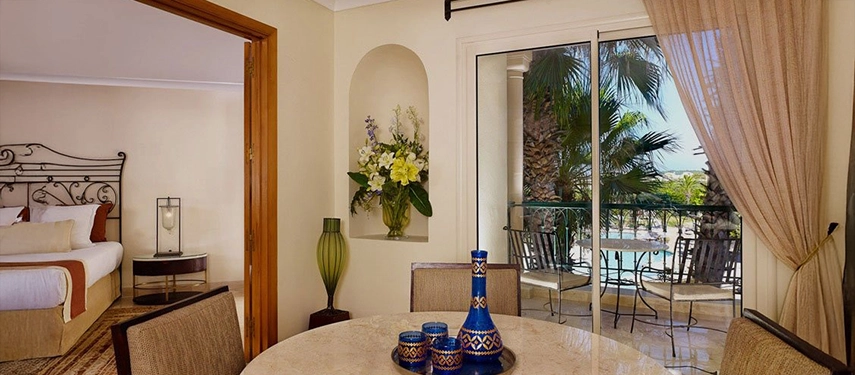 Elegant suite at a Tunisian resort with dining area, floral arrangements, and balcony overlooking palm trees and a pool.