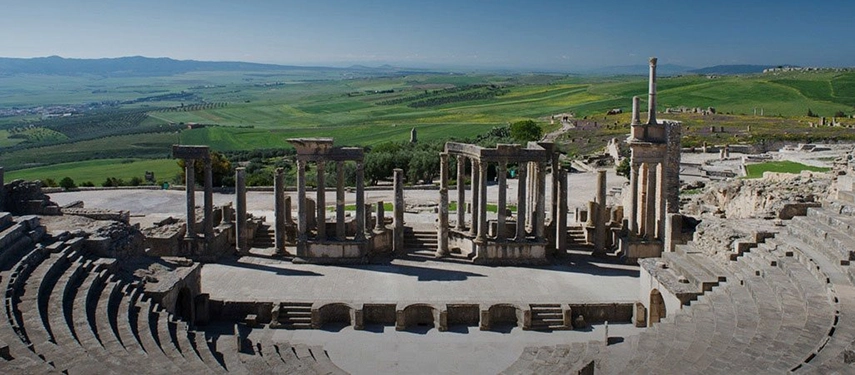 Panoramic view of ancient Roman ruins in Dougga, Tunisia, surrounded by rolling green hills and countryside.