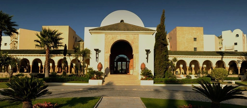 Grand domed entrance to The Residence Tunis, framed by palm trees and arched colonnades under a clear blue sky.