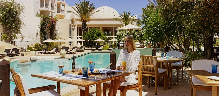Woman enjoying an al fresco lunch by the pool at a restaurant, surrounded by tropical gardens and domed architecture.
