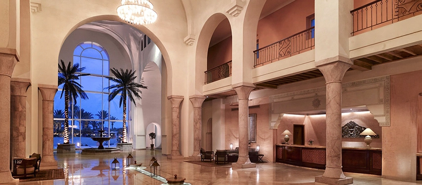 Majestic hotel lobby with marble columns, high ceilings, and arched windows looking onto an illuminated courtyard with palms.