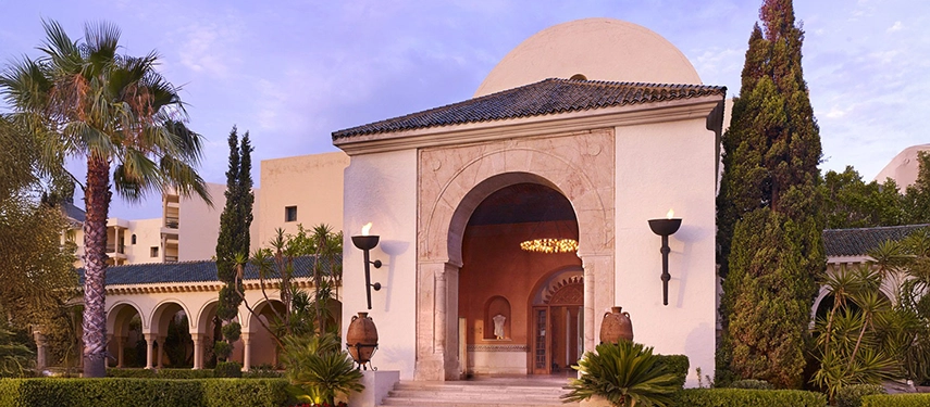 Arched stone entrance of a luxury Tunisian resort at twilight, with lanterns lit and manicured gardens on either side.