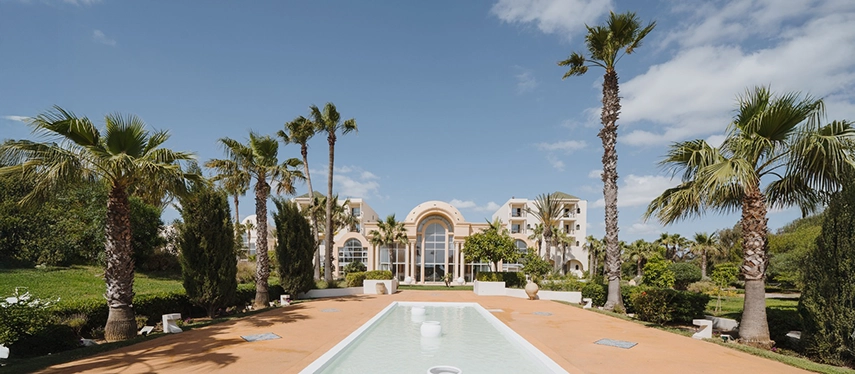 Majestic facade of The Residence Tunis in Tunisia framed by tall palm trees, manicured gardens, and a central reflecting pool.