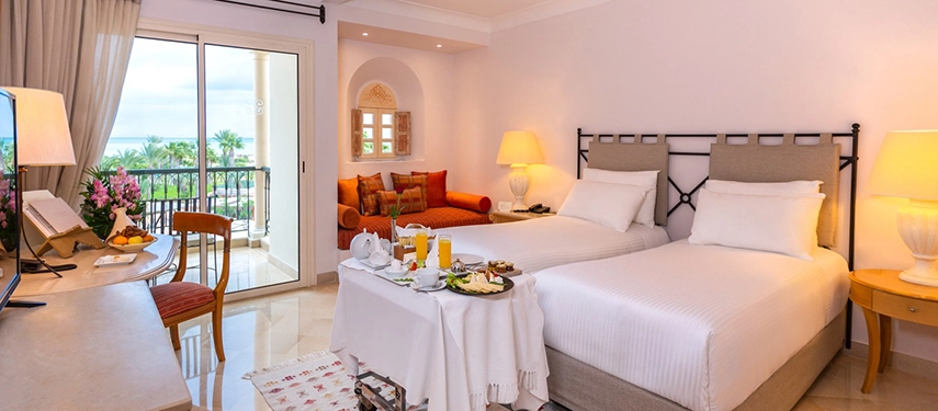 Bright and airy hotel room with twin beds, a breakfast table set for two, and a balcony facing the Mediterranean garden.