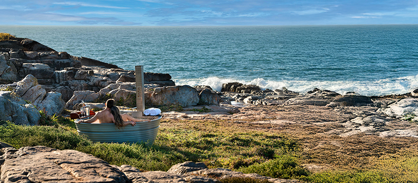 Person relaxing in an outdoor hot tub overlooking the rocky coastline and ocean.