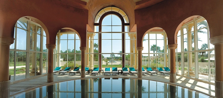 Elegant indoor pool surrounded by arched windows and sun loungers, looking out onto a landscaped Tunisian garden.