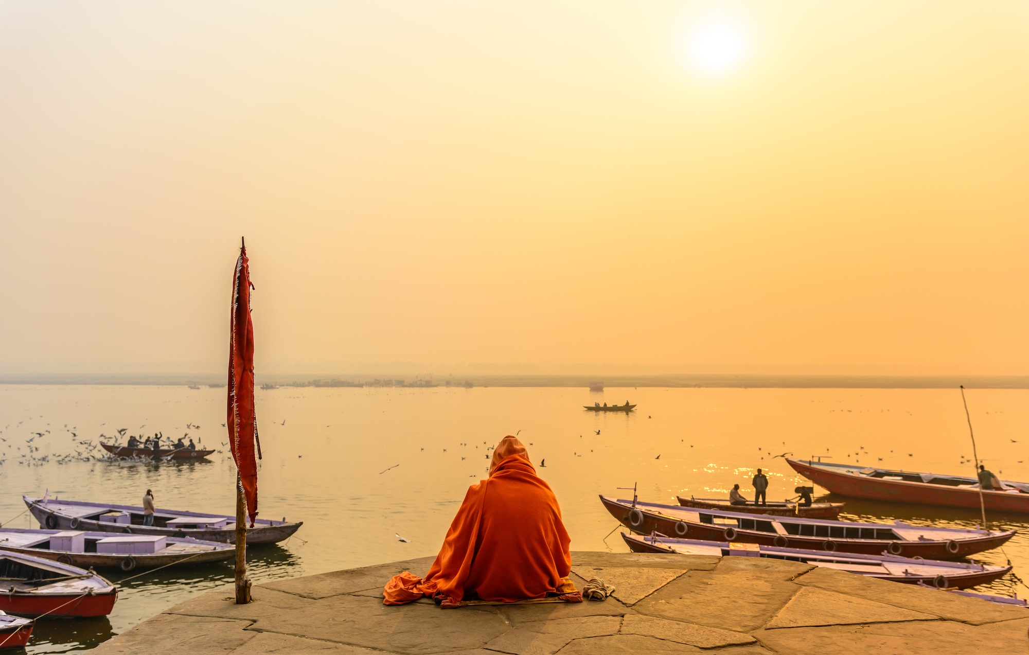 Woman in an orange sari sitting on the ghats of Varanasi, overlooking the Ganges at sunset.