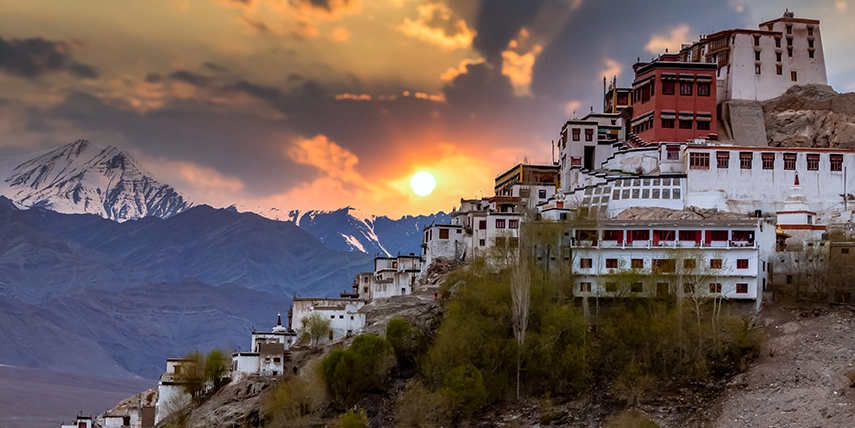 A Himalayan monastery complex perched on a hillside in Ladakh at sunset, overlooking a stark mountain landscape bathed in golden light.