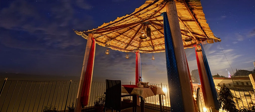 Romantic rooftop dining setup under a thatched canopy overlooking the Ganges River at night.