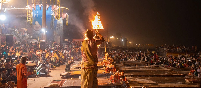 Evening Ganga aarti ceremony with priests holding large flaming oil lamps before a gathered crowd in Varanasi.