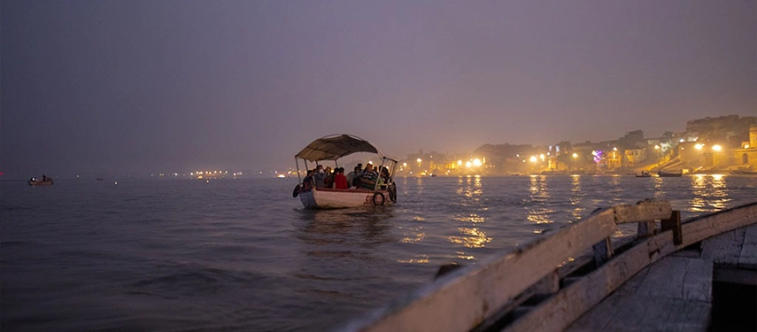 Boat carrying passengers across the Ganges at night with the Varanasi ghats softly illuminated in the distance.