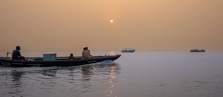 Small wooden boat gliding on the Ganges at sunrise with the sun low on the horizon and other boats in the background.