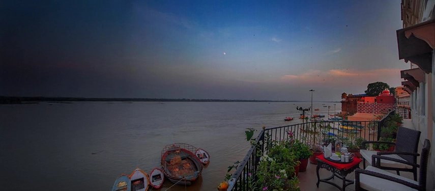 Balcony view over the Ganges River at dusk with moored boats and potted plants along the railing.
