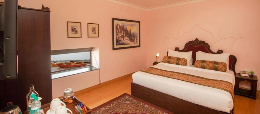 Double room with pastel pink walls, lotus-patterned headboard, and a window view of boats on the Ganges.