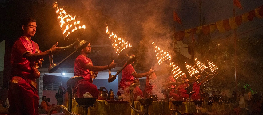 Priests in red robes performing the evening Ganga aarti, each holding large multi-tiered flaming oil lamps.