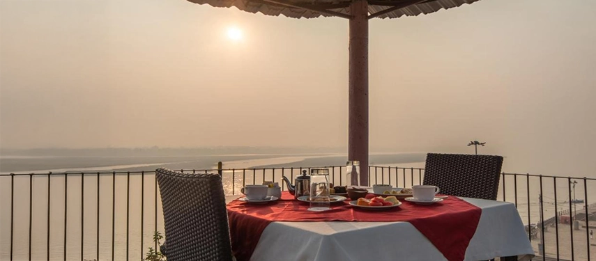 Breakfast table for two under a thatched roof, overlooking the Ganges River at sunrise.