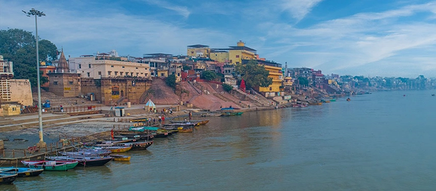 Colourful ghats of Varanasi lining the Ganges River under a clear blue sky with wooden boats moored along the shore.