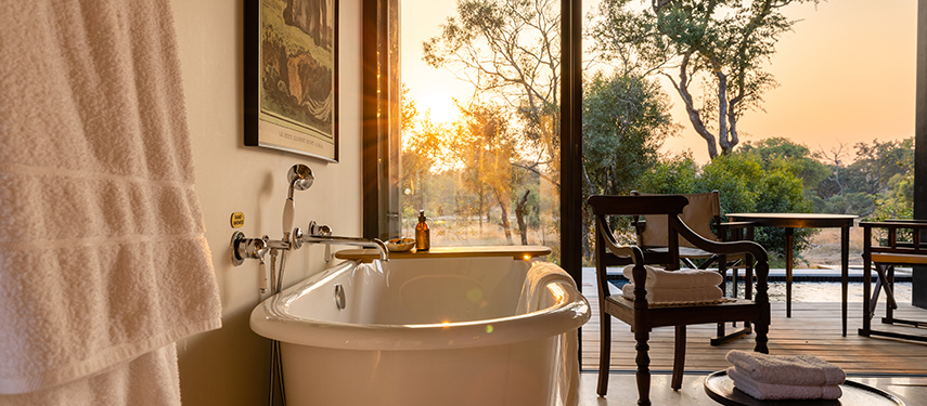 Freestanding bathtub positioned beside a glass wall, overlooking the bushveld as the sun sets.