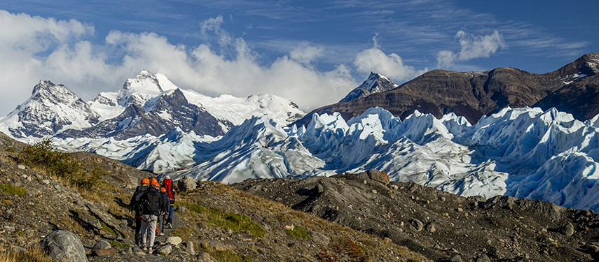 A group of hikers approaches jagged glacier formations beneath towering Andean peaks under a crisp Patagonian sky.