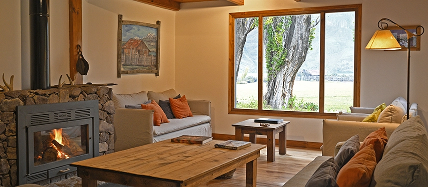 Cozy lounge with sofas, a stone fireplace, and large window views of the Patagonian countryside at Estancia El Cóndor.