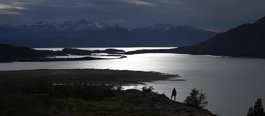 Person silhouetted against the shimmering waters of Lago San Martín under a moody evening sky.